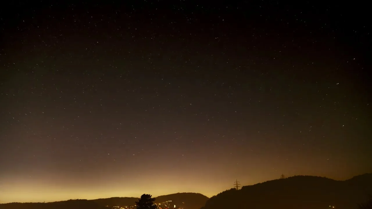 Sternenhimmel über einer Hügelkette, am Horizont das Licht einer Stadt